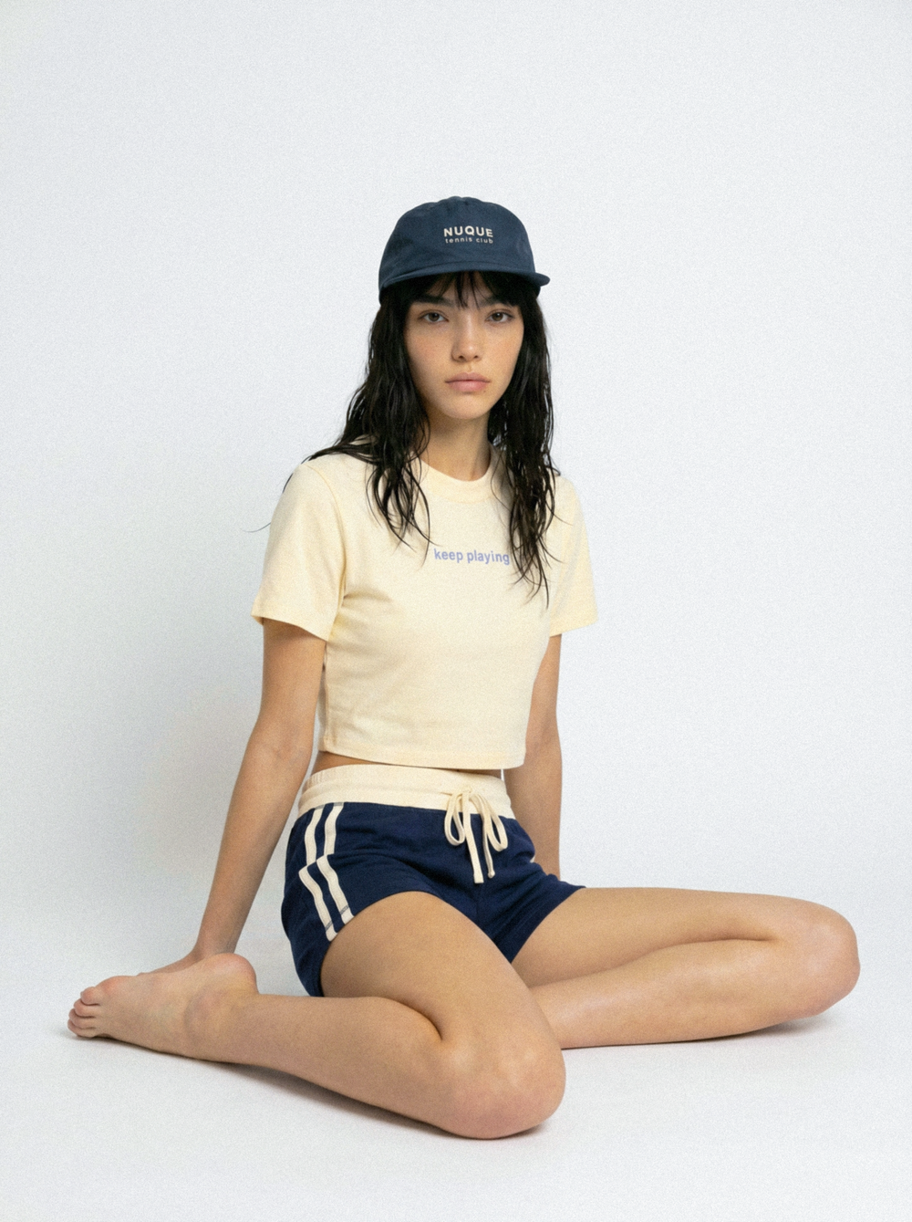 Model wearing a cream Nuque ‘keep playing’ cropped tee, navy retro track shorts with white stripes, and a navy Nuque Tennis Club cap in a minimalist studio setting.
